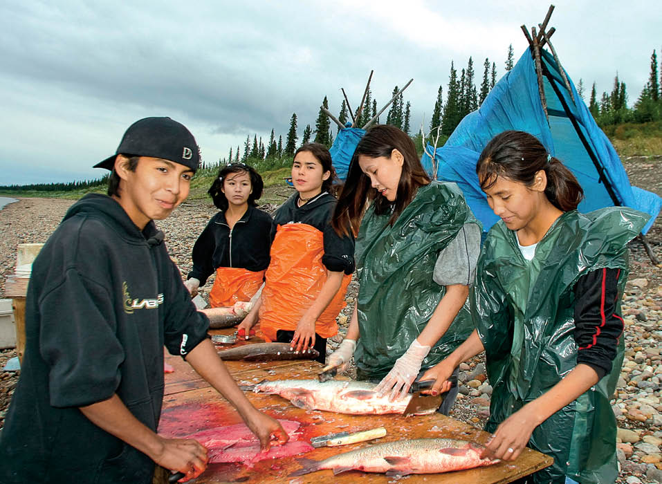 People gathered at shoreline with teepees in the background while gutting fish on a table