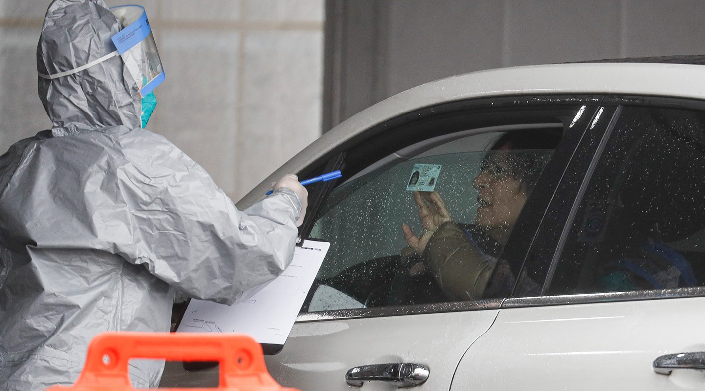 a man driving a car shows ID to a healthcare worker in a hazmat suit